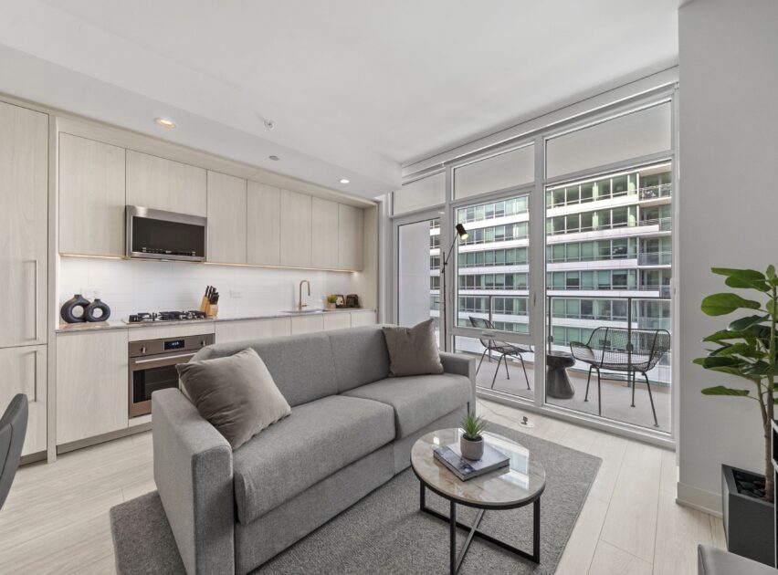 Modern apartment with an open kitchen and living area, featuring light wood cabinets, a grey sofa, a small round coffee table, and large floor-to-ceiling windows leading to a balcony with patio furniture.