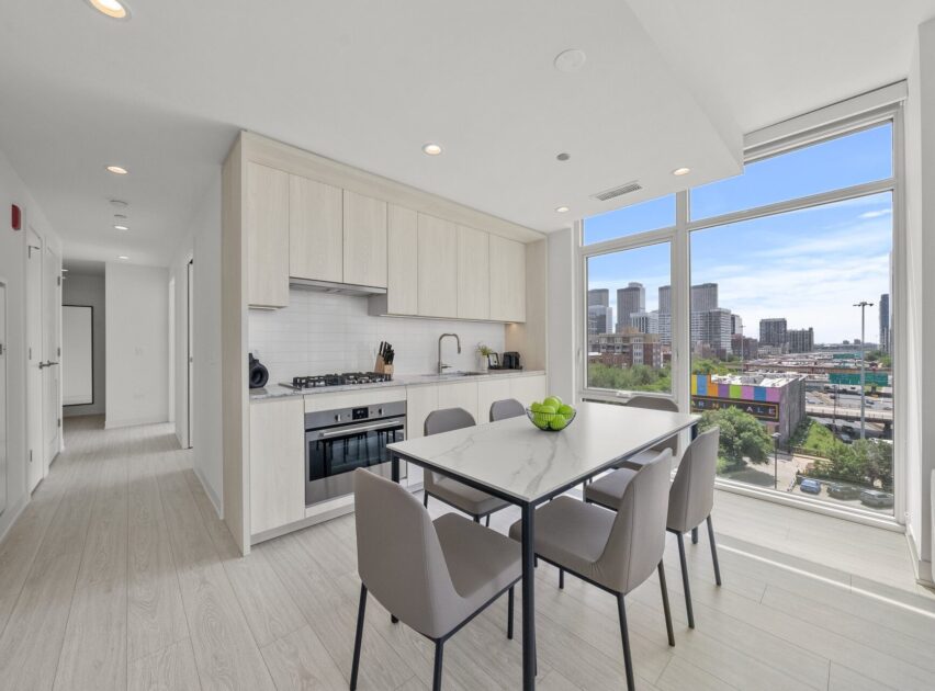 Modern kitchen and dining area with light wood cabinets, built-in appliances, a marble dining table with four chairs, large windows with city views, and plenty of natural light.