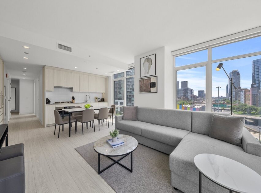 Modern open-concept living room and kitchen with light wood flooring, gray sectional sofa, dining table, and large windows offering a city view with tall buildings and natural light.