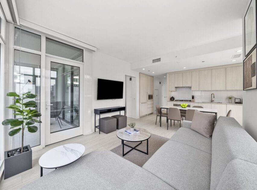 Modern, bright open-concept living room and kitchen with light wood cabinets, gray sectional sofa, round coffee tables, wall-mounted TV, potted plant, and glass doors leading to a balcony.