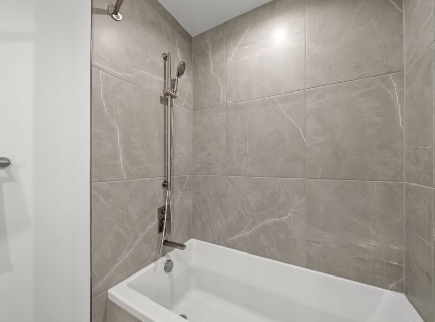 Modern bathroom with a white bathtub, gray marble wall tiles, and a chrome showerhead with a sliding bar mounted above the tub.