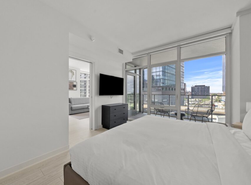 Modern bedroom with large windows, a balcony with outdoor seating, and city views. A bed with white linens, a black dresser, and a wall-mounted TV are visible. An open doorway leads to a bright living area.