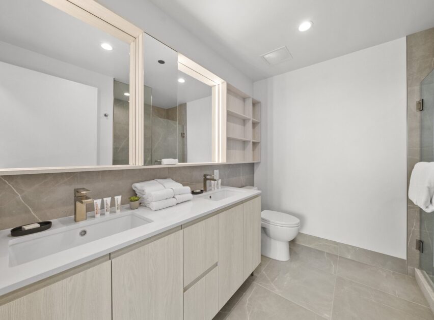 Modern bathroom with light wood double vanity, large mirrors, neatly folded towels, toiletries, a toilet, open wall shelves, and a glass-enclosed shower; neutral gray and white color scheme, bright lighting.