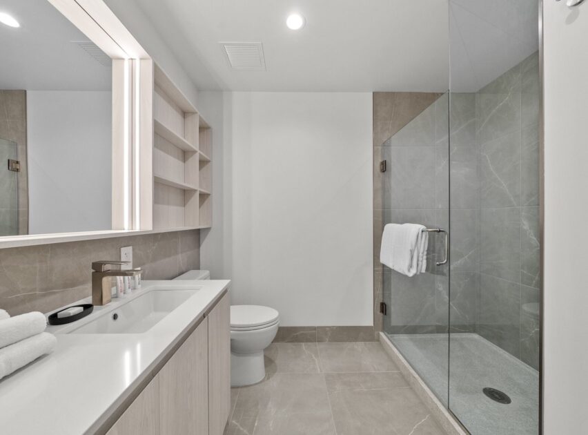 Modern bathroom with light-colored tile flooring, a glass-enclosed shower with a white towel on the handle, a wall-mounted toilet, a large vanity with double sinks, and open shelving next to a wide mirror.