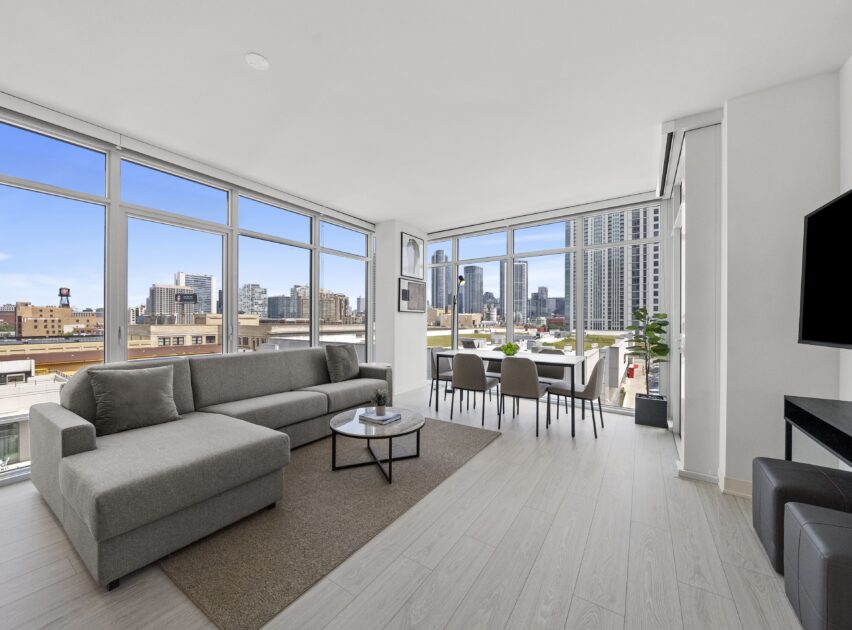 A modern, bright living and dining area with large floor-to-ceiling windows, gray sectional sofa, dining table, and city skyline views. Light wood floors and minimalistic decor create an open, airy atmosphere.