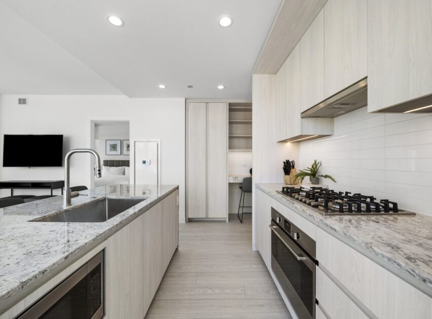 Modern kitchen with light wood cabinets, a marble countertop island with sink, built-in ovens, a gas stovetop, and a workspace nook in the background. The space is bright with recessed lighting and minimal decor.