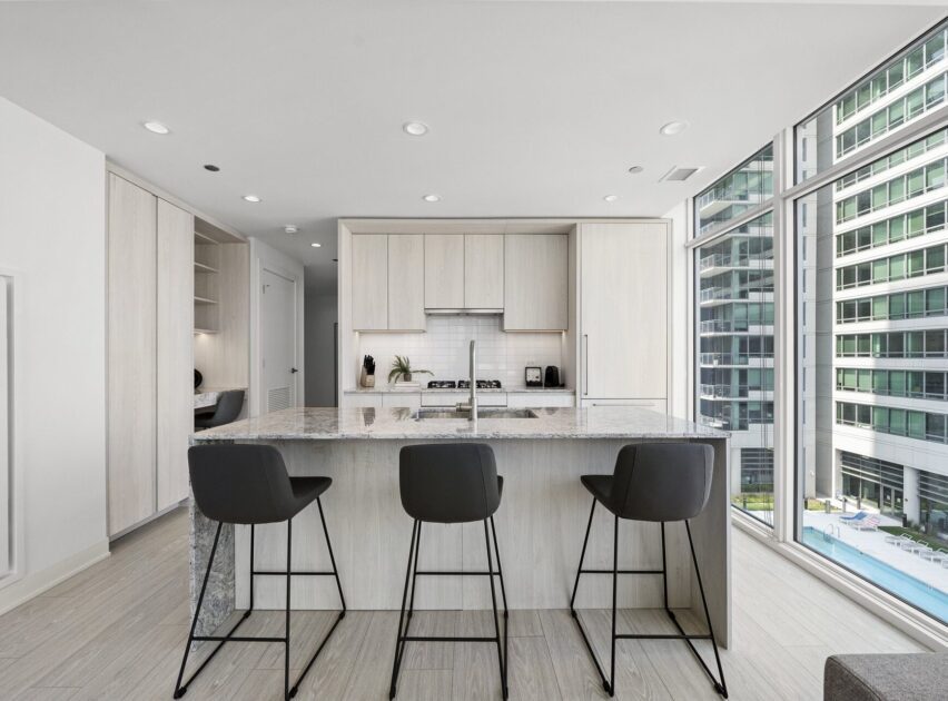 Modern kitchen with light wood cabinets, a marble island countertop, and three black barstools. Large floor-to-ceiling windows provide a view of nearby buildings and a swimming pool outside.