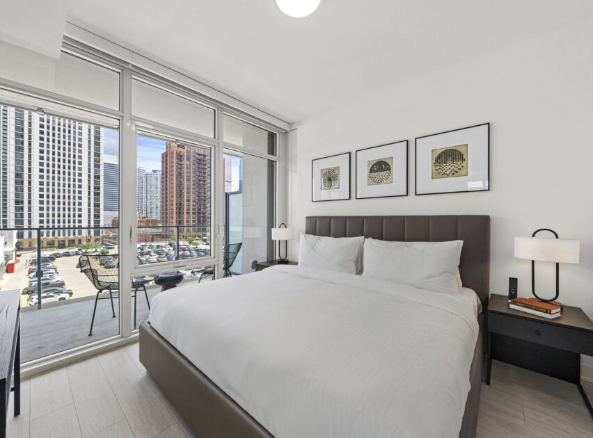 Modern bedroom with a large bed, two nightstands, artwork on the wall, floor-to-ceiling windows, and a balcony overlooking a cityscape with high-rise buildings and a parking lot.