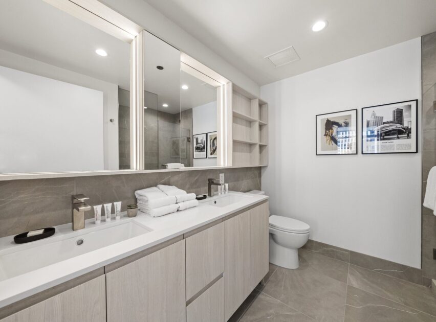 Modern bathroom with light wood cabinetry, double sinks, stacked towels, toiletries, a toilet, wall art, open shelving, and large mirrors. The room has gray tiled flooring and neutral-toned walls.