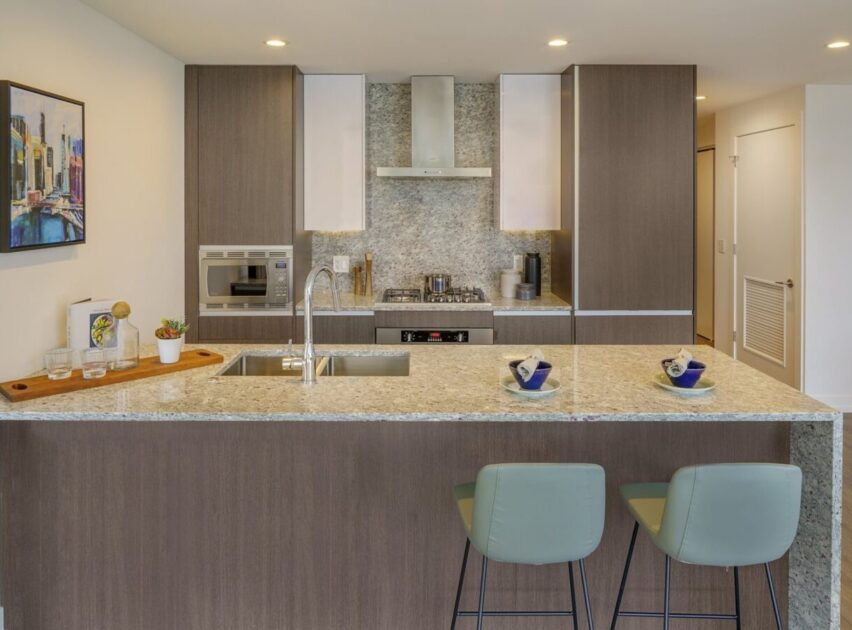 Modern kitchen with a marble island, two light green stools, built-in oven, stove, stainless steel range hood, sink, and minimalist decor. Wall art and neatly arranged utensils add a contemporary touch.
