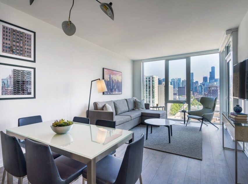 Modern apartment living room with a gray sofa, coffee tables, dining table with chairs, large windows with city skyline view, artwork on white walls, and natural light filling the space.
