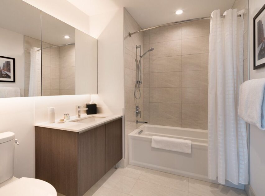 Modern bathroom with a wooden vanity, white countertop, wall-mounted mirror cabinets, toilet, bathtub with a shower, beige tiled wall, white shower curtain, towel rack, and framed photo on the wall.
