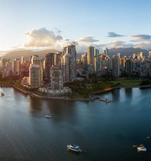 Aerial view of Vancouver skyline with high-rise buildings along the waterfront, boats in the water, green parks, and mountains in the background under a partly cloudy sky.