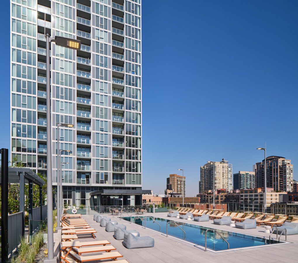 A modern rooftop pool area with lounge chairs and cabanas sits in front of a tall glass apartment building, with city skyscrapers visible under a clear blue sky.