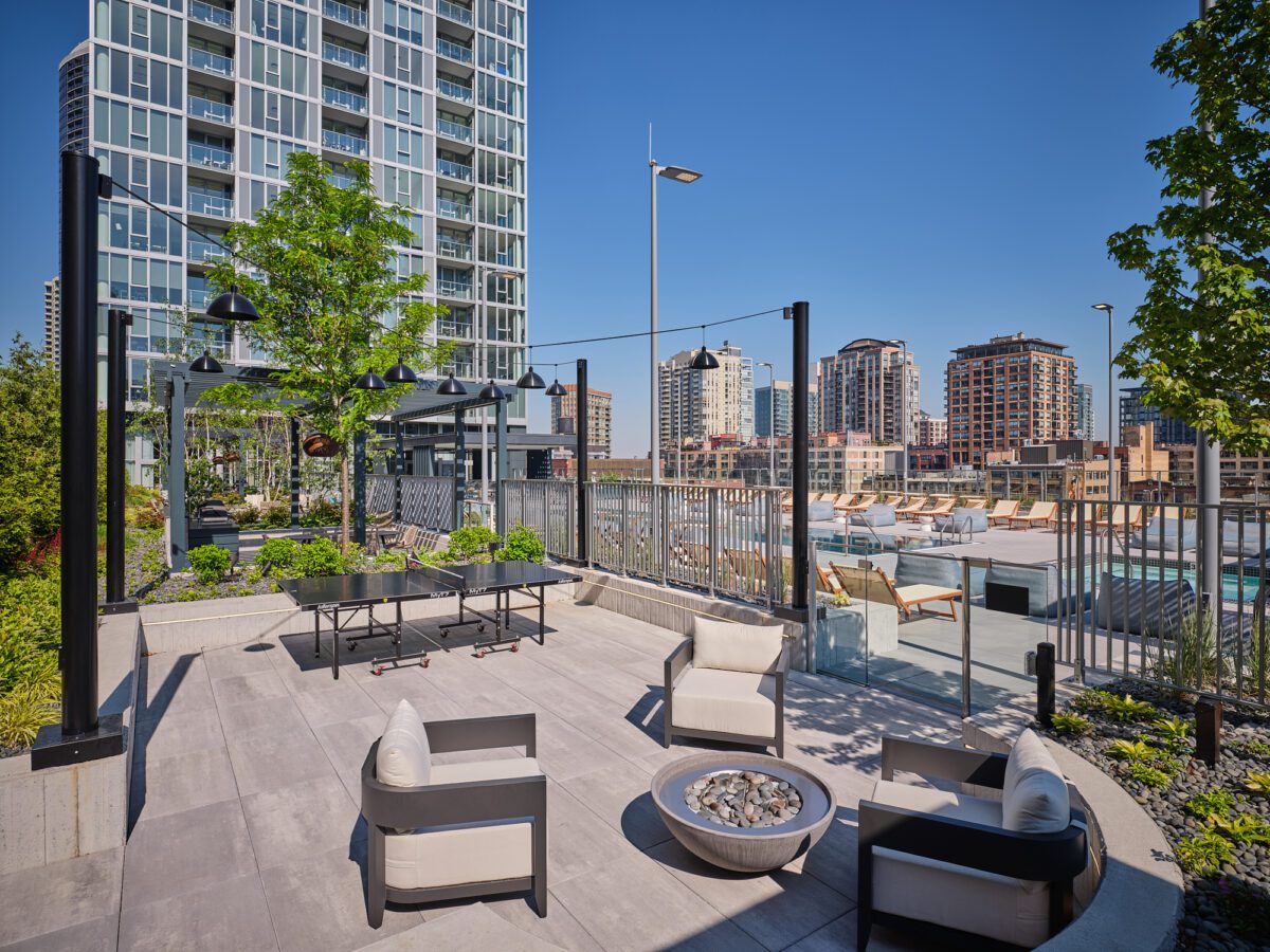 Outdoor rooftop terrace with modern seating, a fire pit, and a ping pong table, overlooking a cityscape with high-rise buildings and a pool area under a clear blue sky.