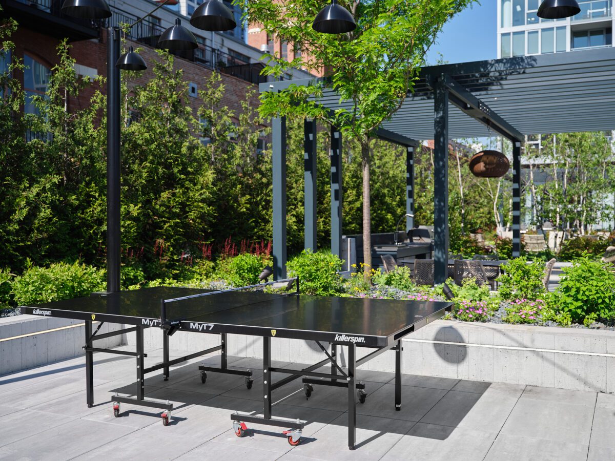A black outdoor ping pong table on a rooftop terrace surrounded by greenery, modern pergolas, lounge seating, and overhead lights, with buildings visible in the background.