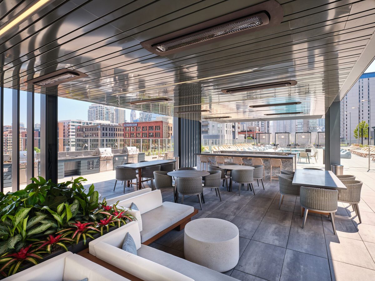 Modern rooftop patio with tables, chairs, couches, and potted plants. The area is covered with a sleek metal roof and surrounded by city buildings, offering an open, urban view and plenty of sunlight.