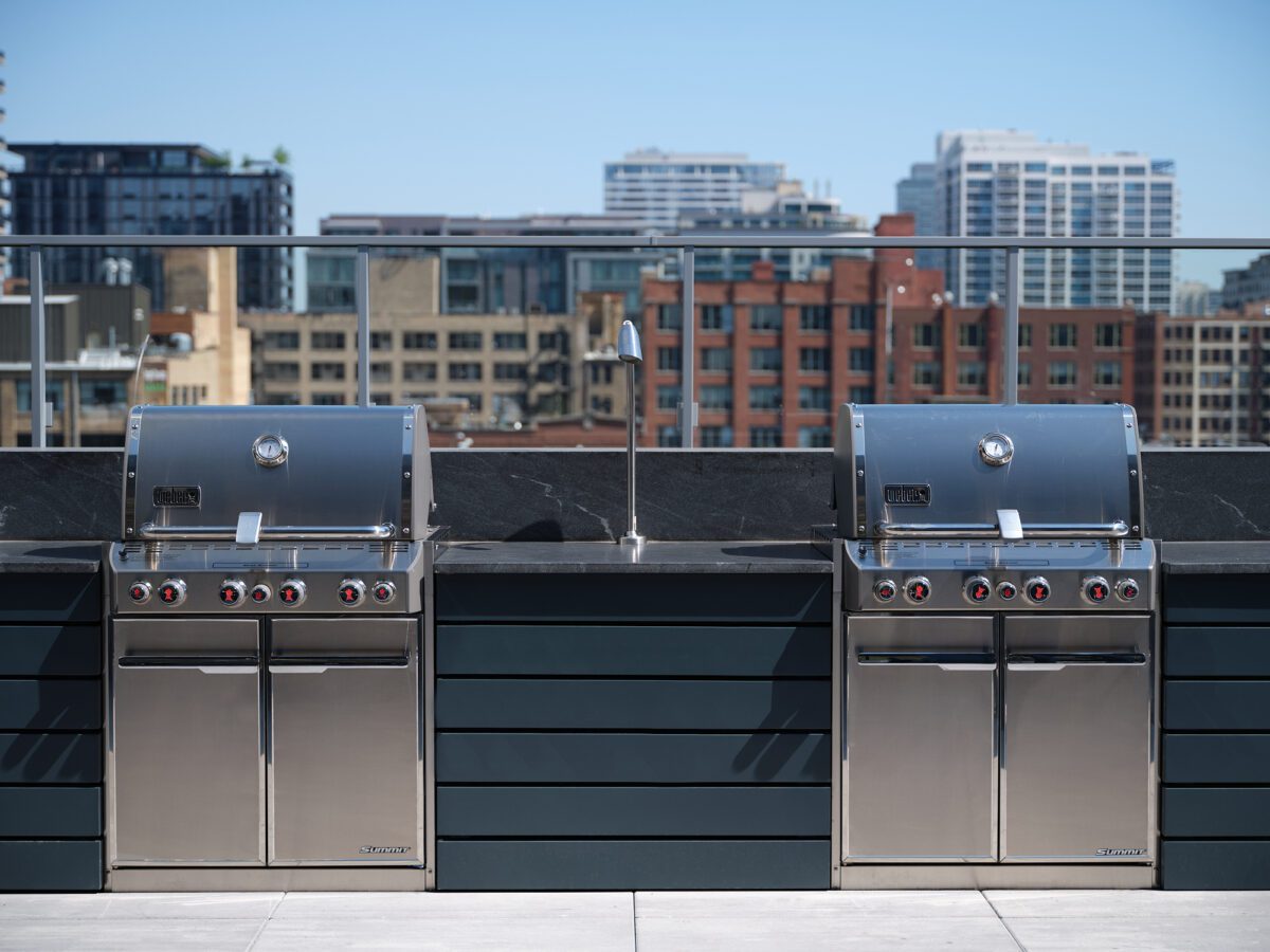 Two stainless steel outdoor grills are side by side on a rooftop patio with a glass railing, overlooking a cityscape with modern buildings under a clear blue sky.