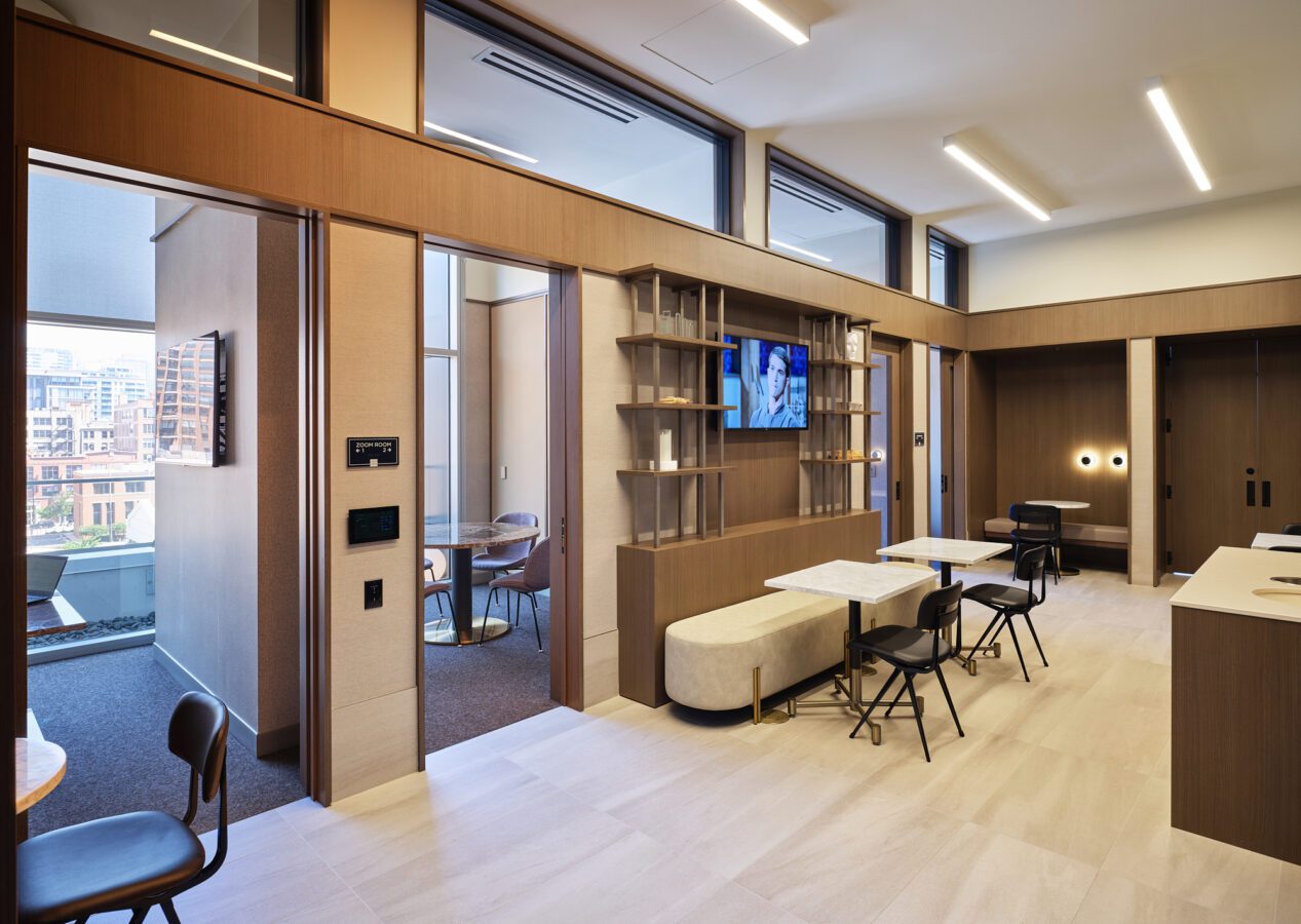 Modern office lounge with light wood finishes, marble tables, black chairs, a TV on the wall, and large windows. Private meeting rooms with glass walls are visible on the left, overlooking a cityscape.