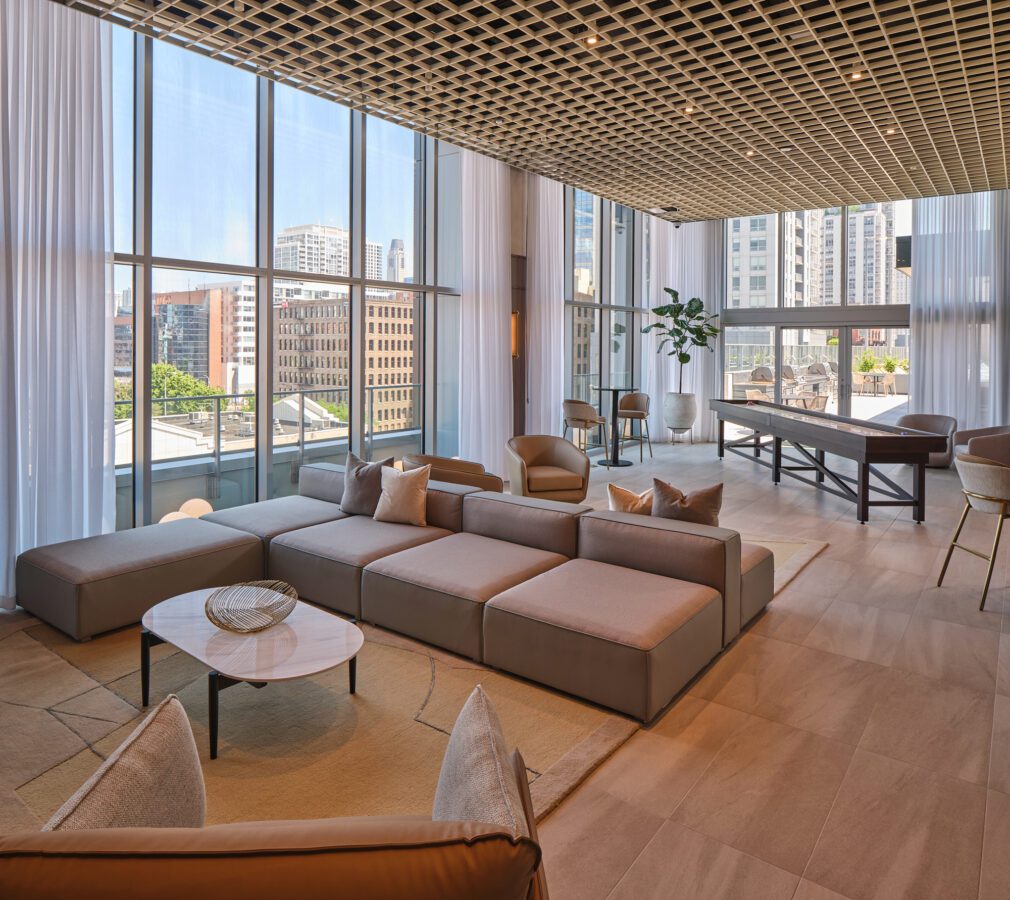 Modern lounge with floor-to-ceiling windows, a large sectional sofa, round coffee table, armchairs, a pool table, bar stools, and city views. Light wood flooring and a grid-patterned ceiling complete the space.