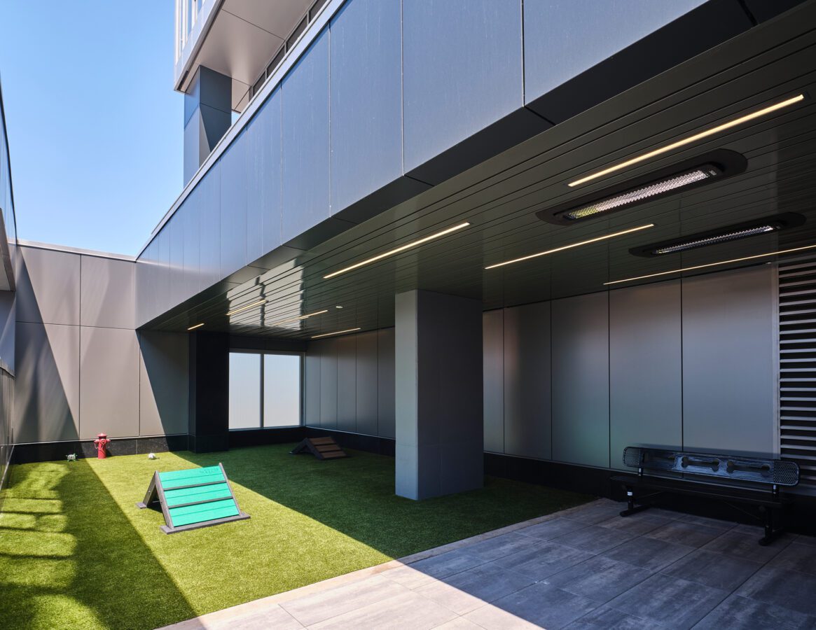 A modern outdoor area with artificial grass, agility ramps for dogs, a bench, and sleek gray building walls, partially covered and lit by linear ceiling lights.
