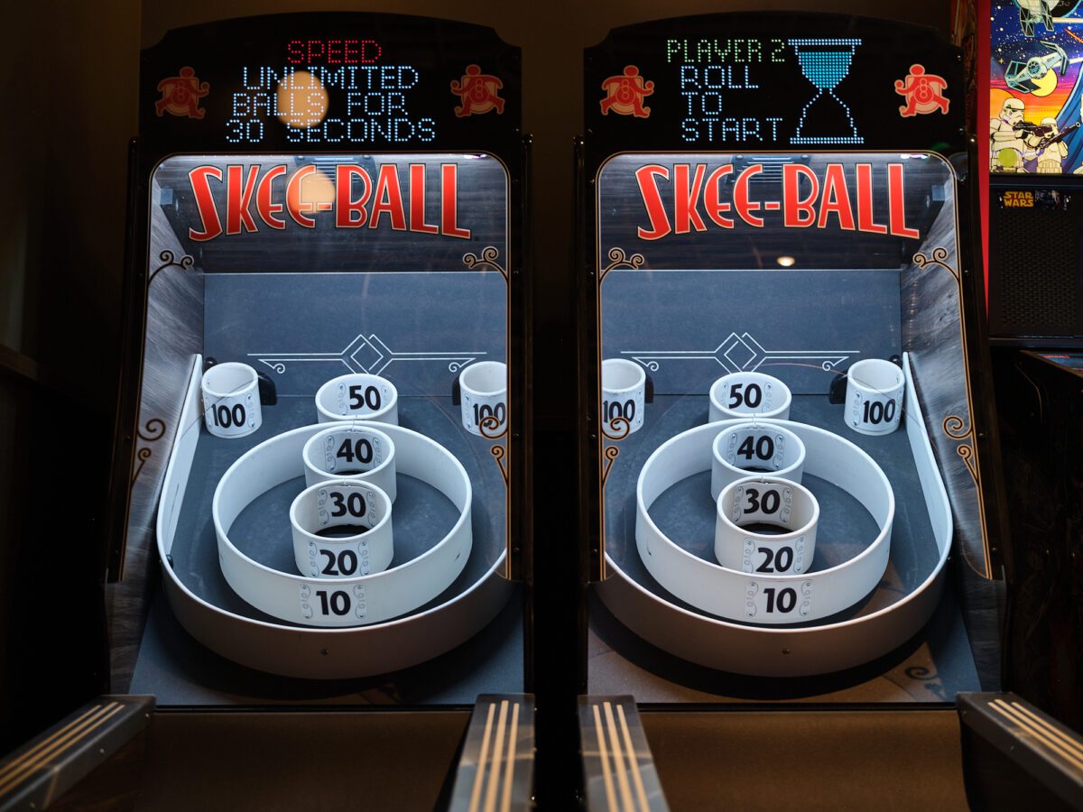 Two Skee-Ball arcade machines side by side, brightly lit. Each machine has scoring rings labeled with points from 10 to 100. Digital displays at the top show instructions for playing.