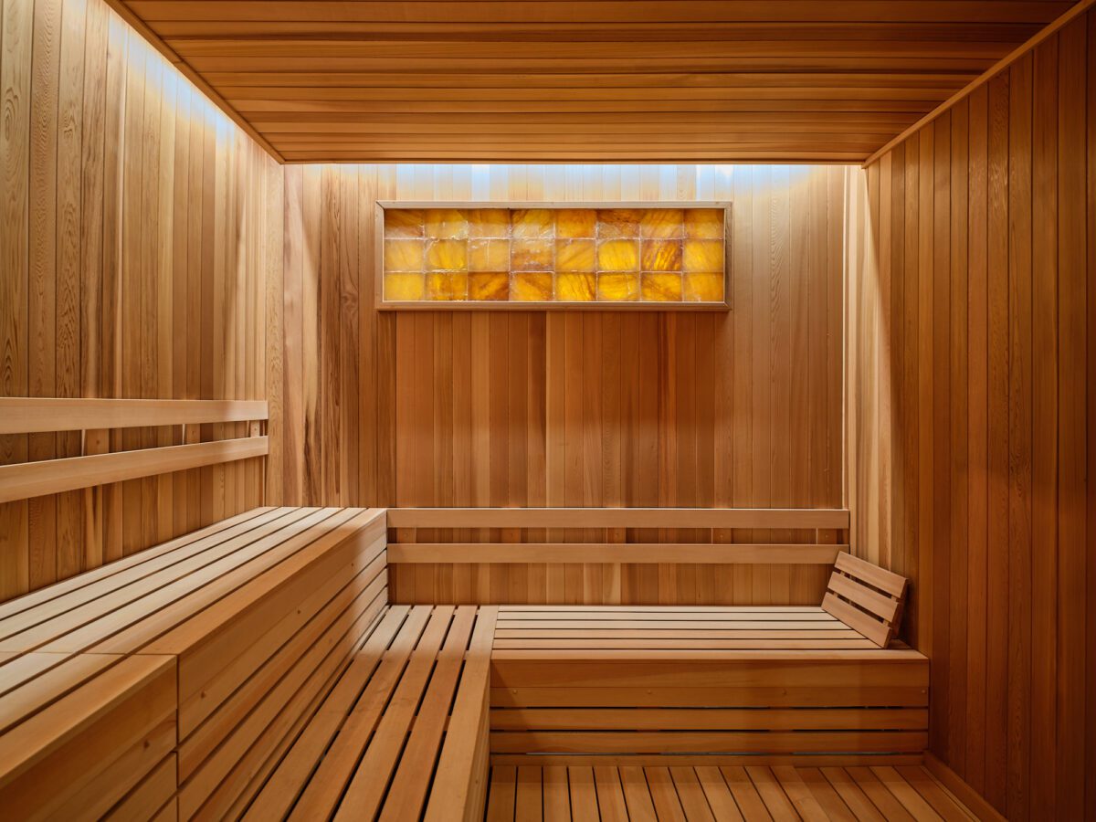 A modern wooden sauna interior with tiered benches, warm overhead lighting, and a decorative backlit panel on the wall. The walls, ceiling, and benches are made of smooth, light-colored wood.