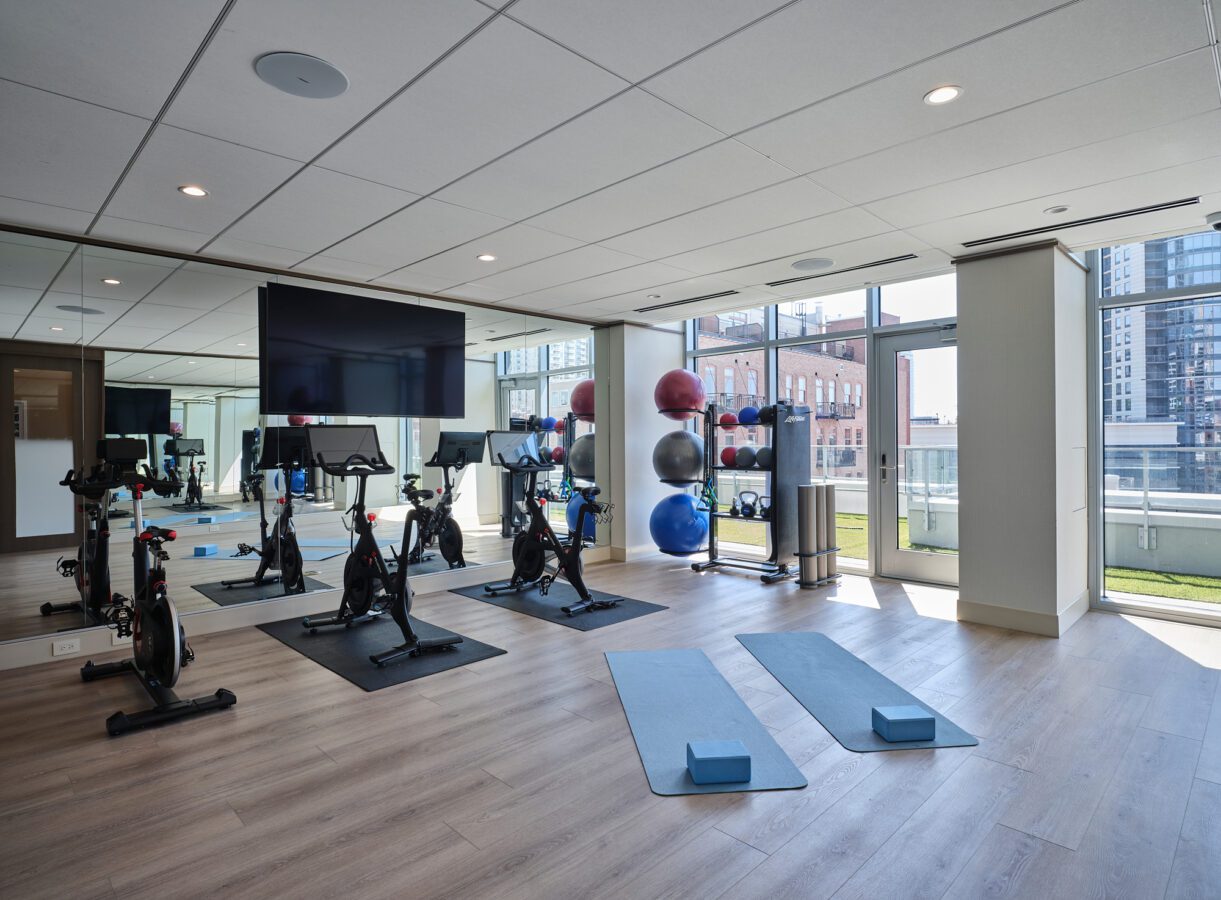 A modern fitness room with stationary bikes, yoga mats, yoga blocks, and exercise balls on racks. Large windows let in natural light and offer a view of city buildings outside.