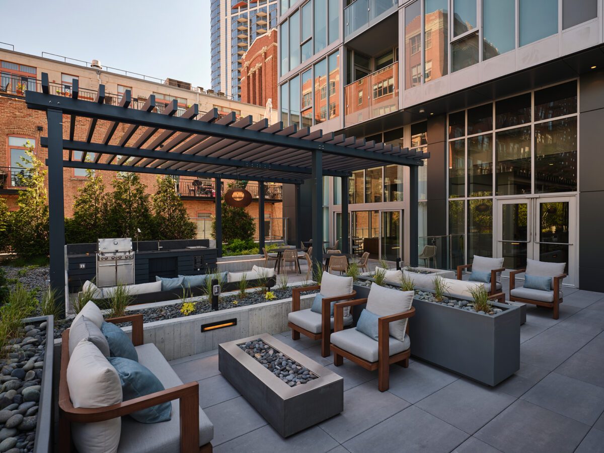 Modern outdoor patio with cushioned seating, a fire pit, and pergola, surrounded by plants and urban buildings. Glass doors and windows reflect the cityscape in the background.