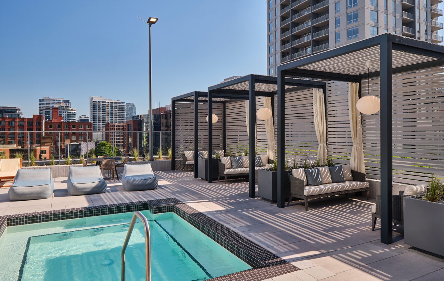 Rooftop pool area with lounge chairs and modern cabanas featuring cushions and hanging lights, set against a city skyline with tall buildings under a clear blue sky.
