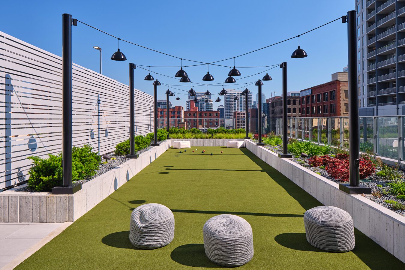 A rooftop bocce ball court with green turf, string lights overhead, three round gray poufs in the foreground, garden beds on the sides, and city buildings in the background under a clear blue sky.