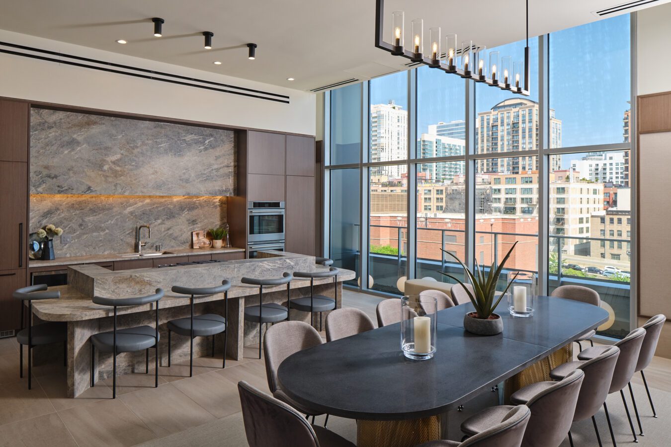 Modern kitchen and dining area with marble countertops, bar stools, a long dining table, floor-to-ceiling windows, and cityscape views in the background. Stylish lighting fixtures hang from the ceiling.
