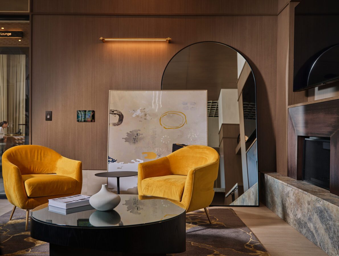 A modern living room with two yellow velvet chairs, a round glass coffee table with books and a vase, abstract art, a large arched mirror, and wood-paneled walls. Warm lighting creates a cozy atmosphere.