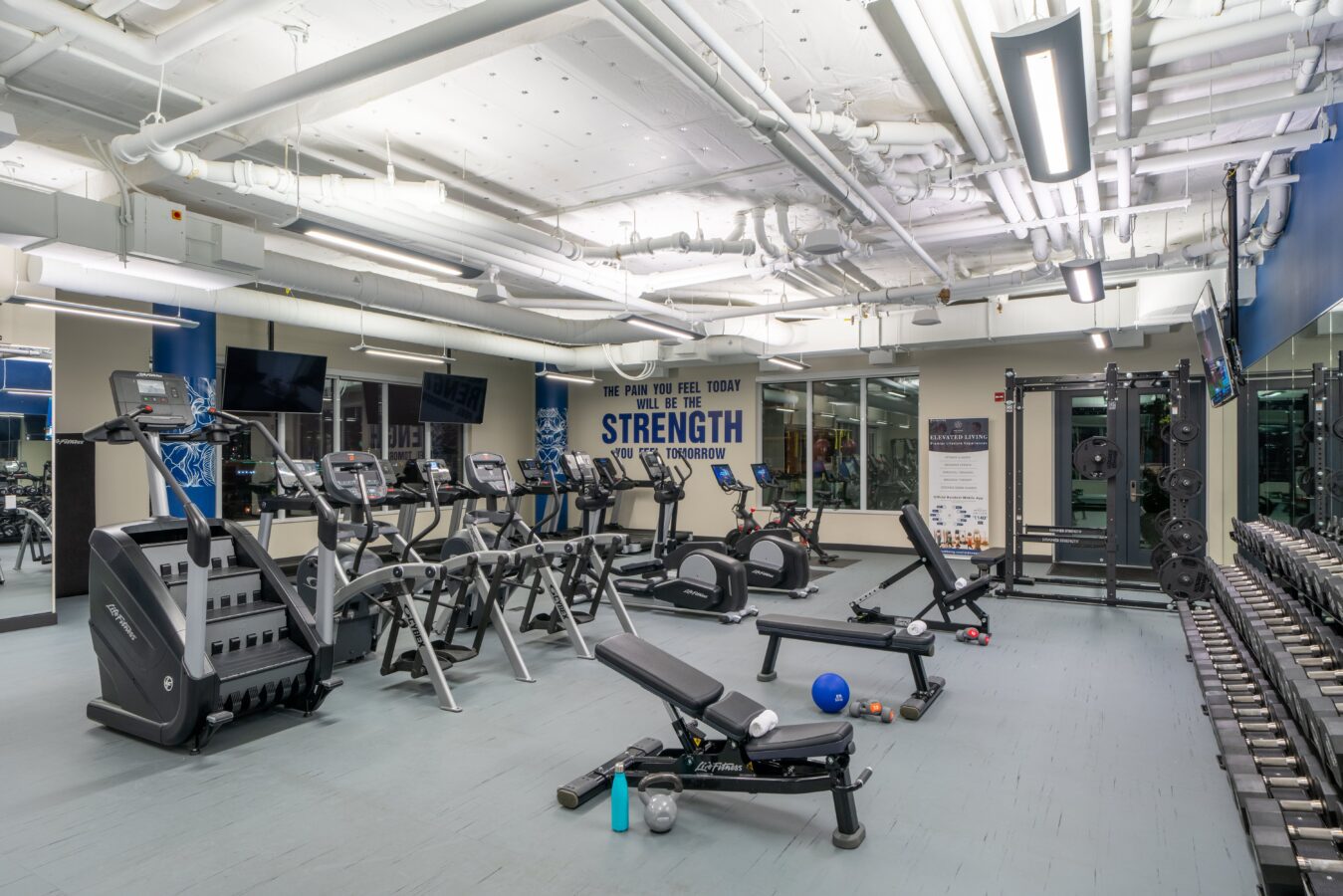 A modern gym with treadmills, ellipticals, stationary bikes, weight benches, free weights, dumbbells, mirrors, and motivational posters on the wall. The area is brightly lit with overhead fluorescent lights.