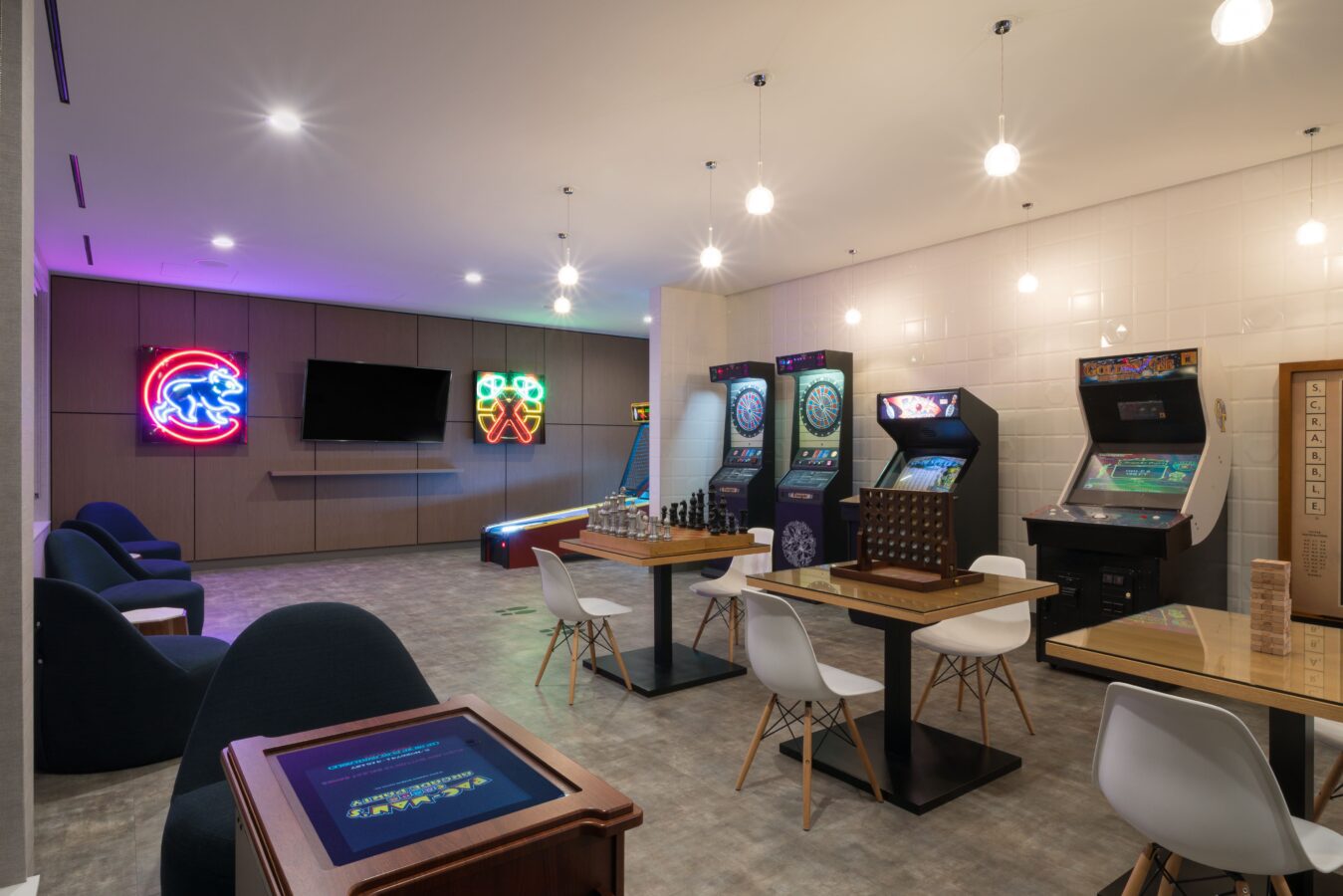 A modern game room with arcade machines, a TV, neon wall signs, tables with chess and board games, and modern chairs under several ceiling lights. The space has a relaxed and playful atmosphere.
