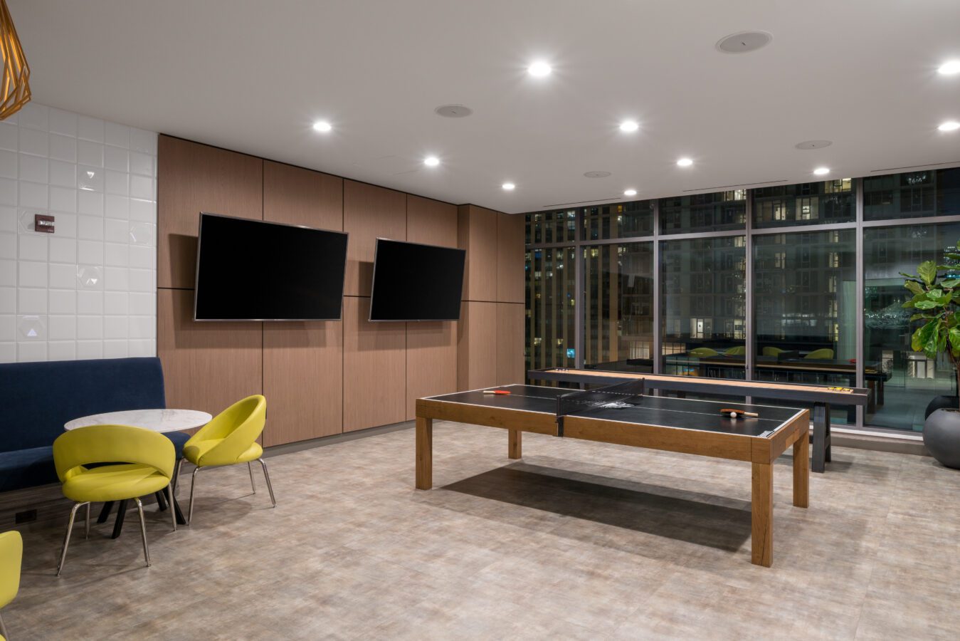 A modern game room with a ping pong table, two wall-mounted flat-screen TVs, a round table with yellow chairs, and large floor-to-ceiling windows overlooking a cityscape at night.