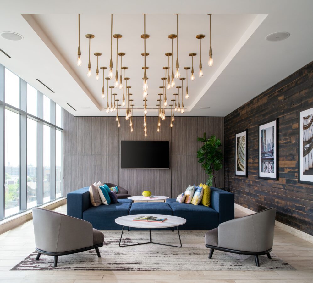 A modern lounge with a blue sectional sofa, two gray chairs, a round coffee table, and colorful pillows. Large windows, a wood accent wall with art, a TV, a potted plant, and hanging pendant lights complete the space.