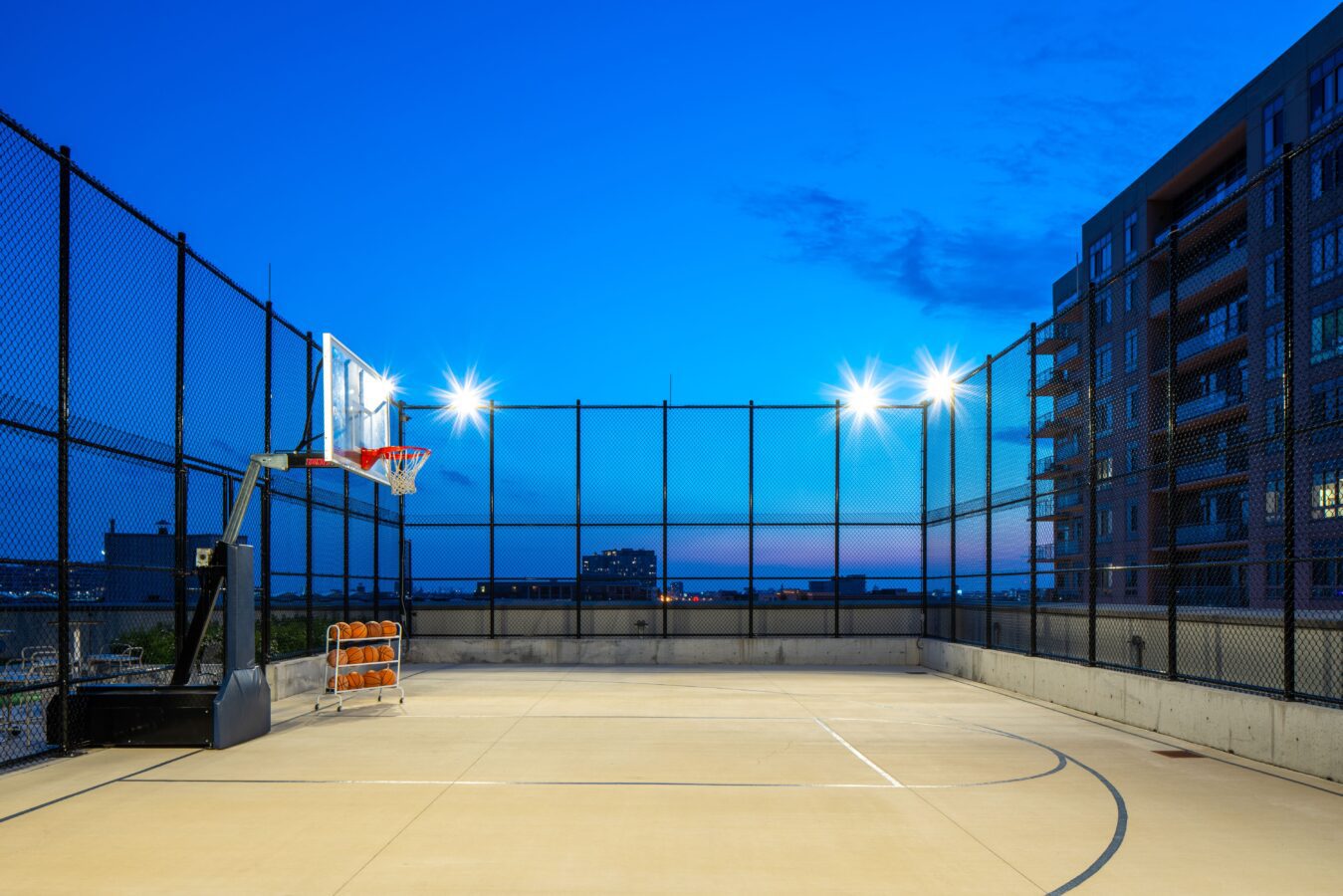 An outdoor basketball court on a rooftop, surrounded by a tall black fence, brightly lit by overhead lights at dusk, with apartment buildings visible on the right and a clear blue sky in the background.