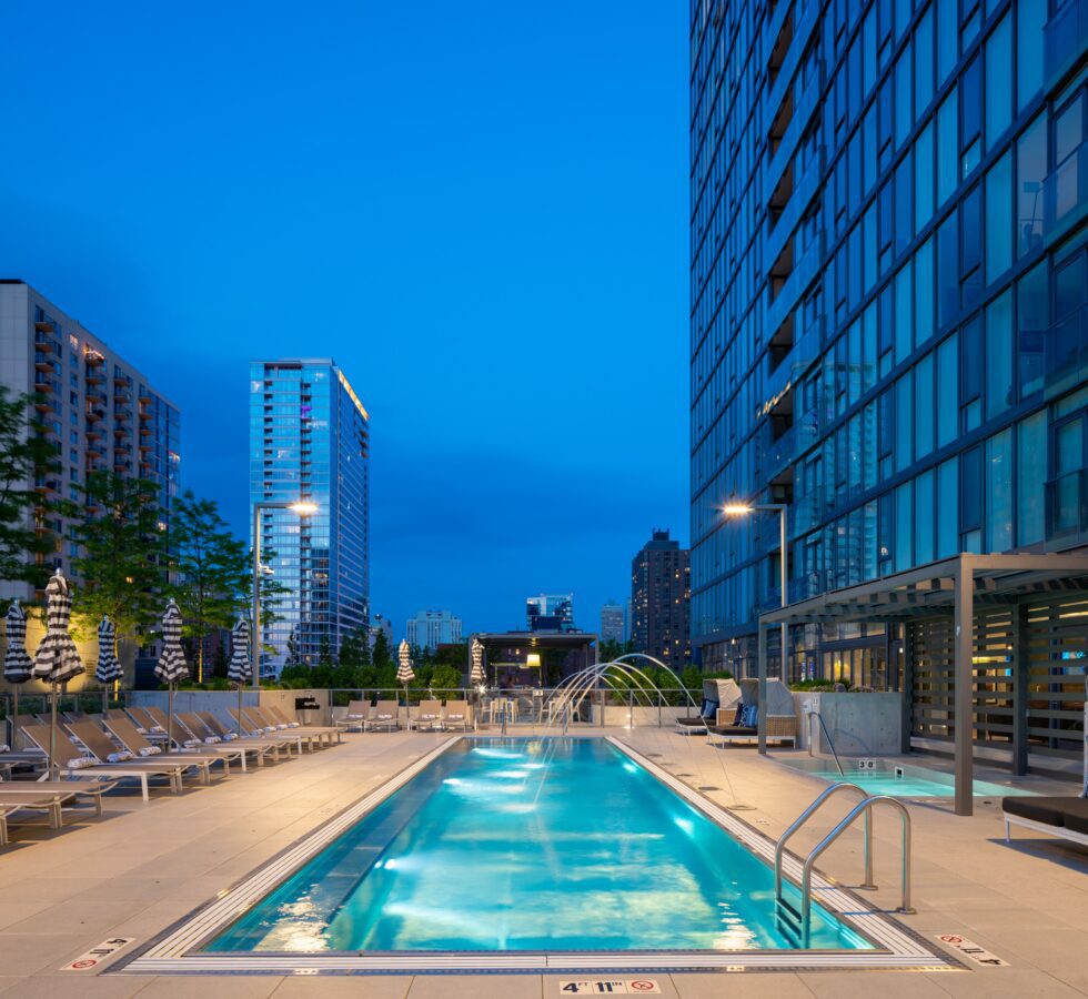 A modern outdoor swimming pool at night, surrounded by lounge chairs and umbrellas, with city skyscrapers and a clear blue sky in the background. The pool area is well-lit and inviting.