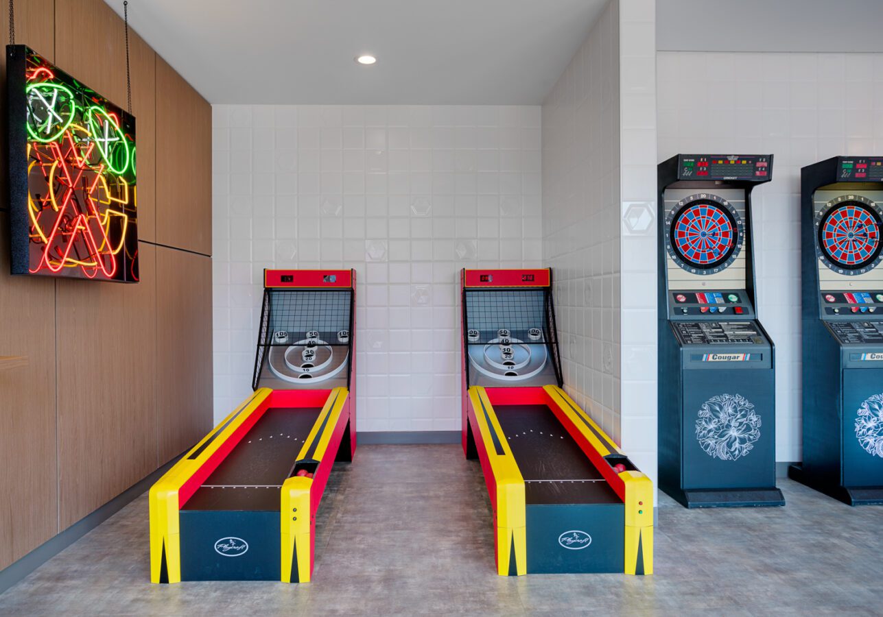 Two brightly colored skee-ball machines and three electronic dartboards are set up in a clean, modern game room with a neon sign on the wall and white tiled accents.