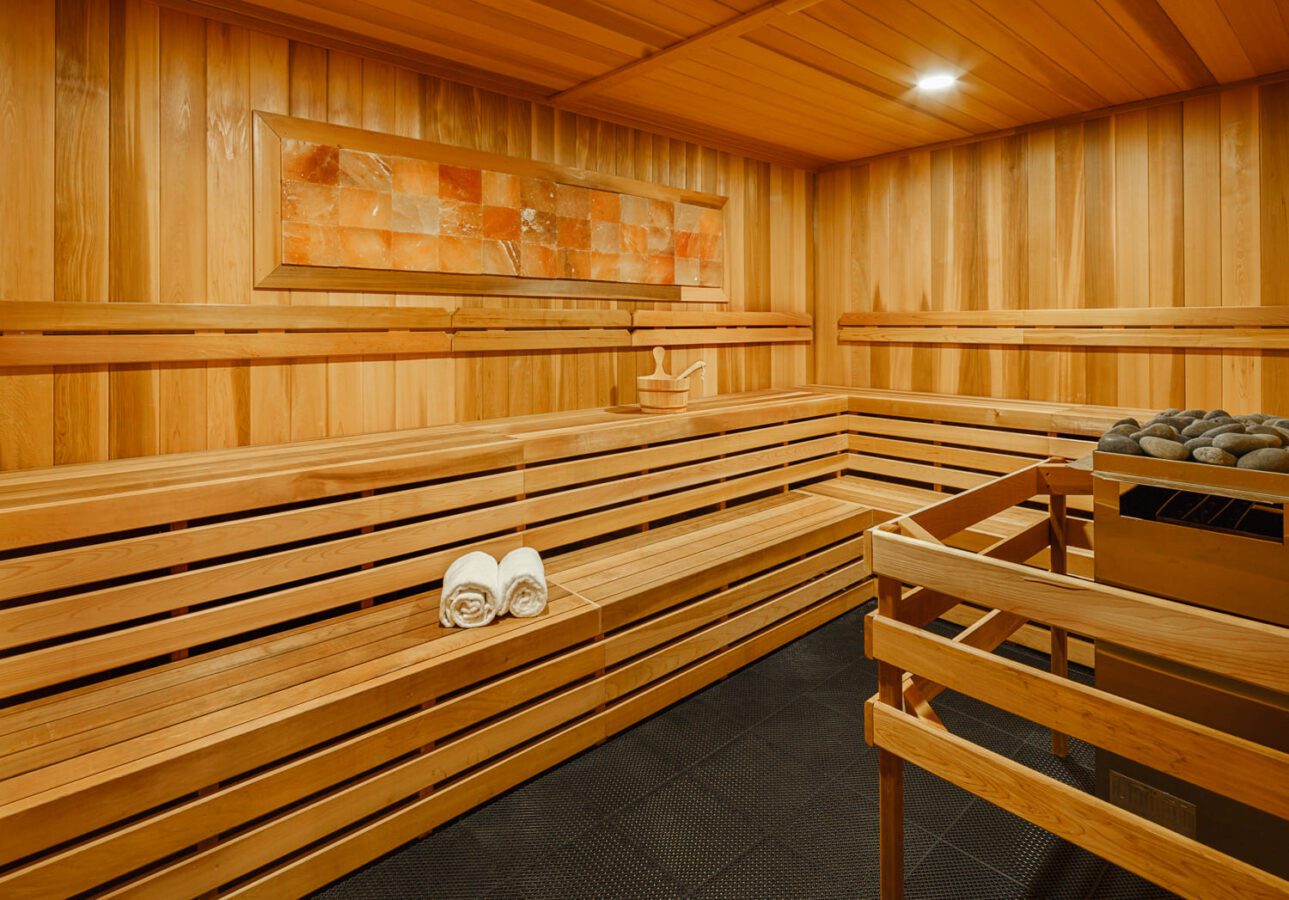 A spacious wooden sauna room with tiered benches, two rolled white towels, a bucket with a ladle, and a heater filled with sauna stones; a salt rock panel decorates the wall, and the lighting is warm.