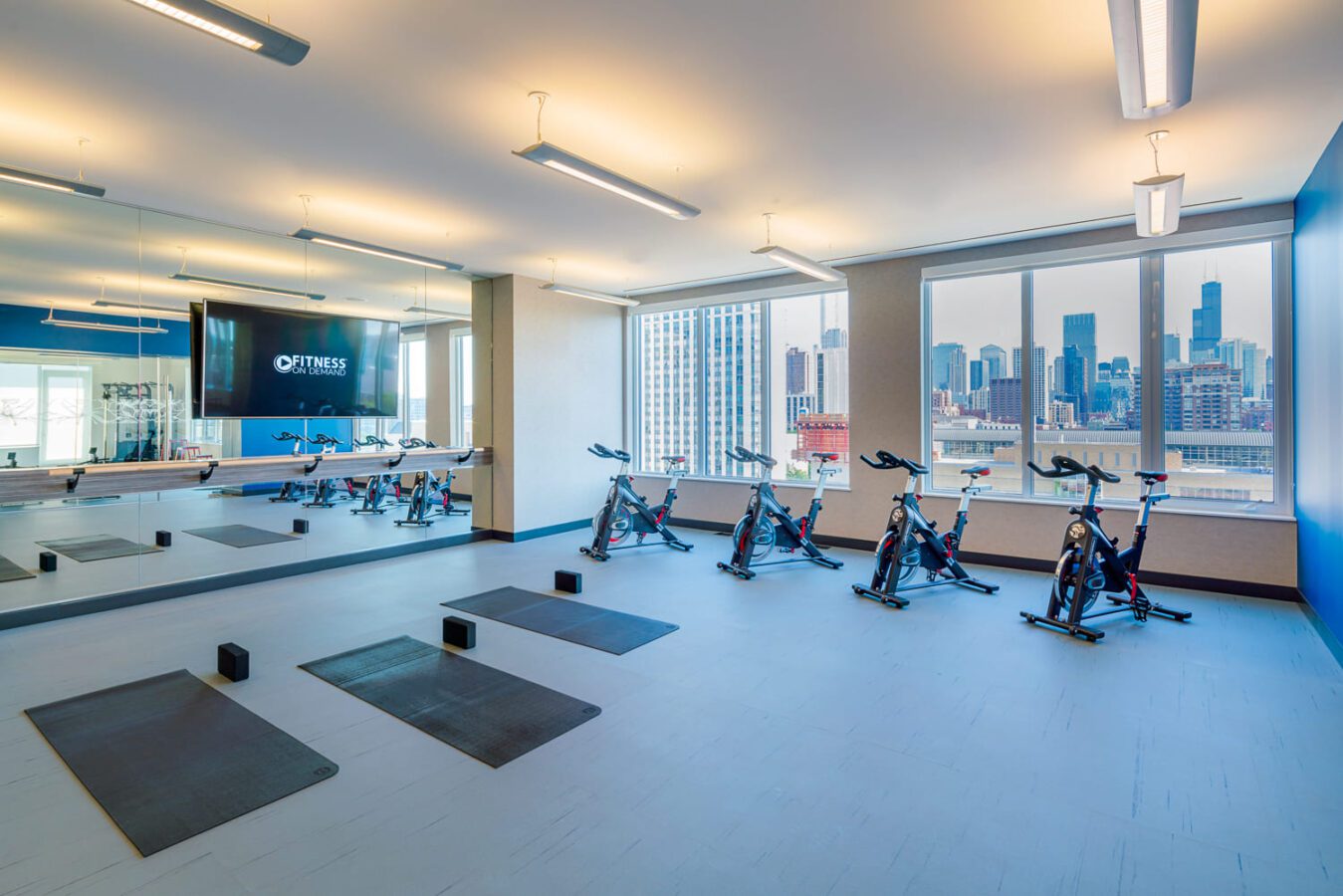 A modern gym studio features floor mats, foam blocks, stationary bikes, large mirrors, and tall windows with a city skyline view under bright ceiling lights.