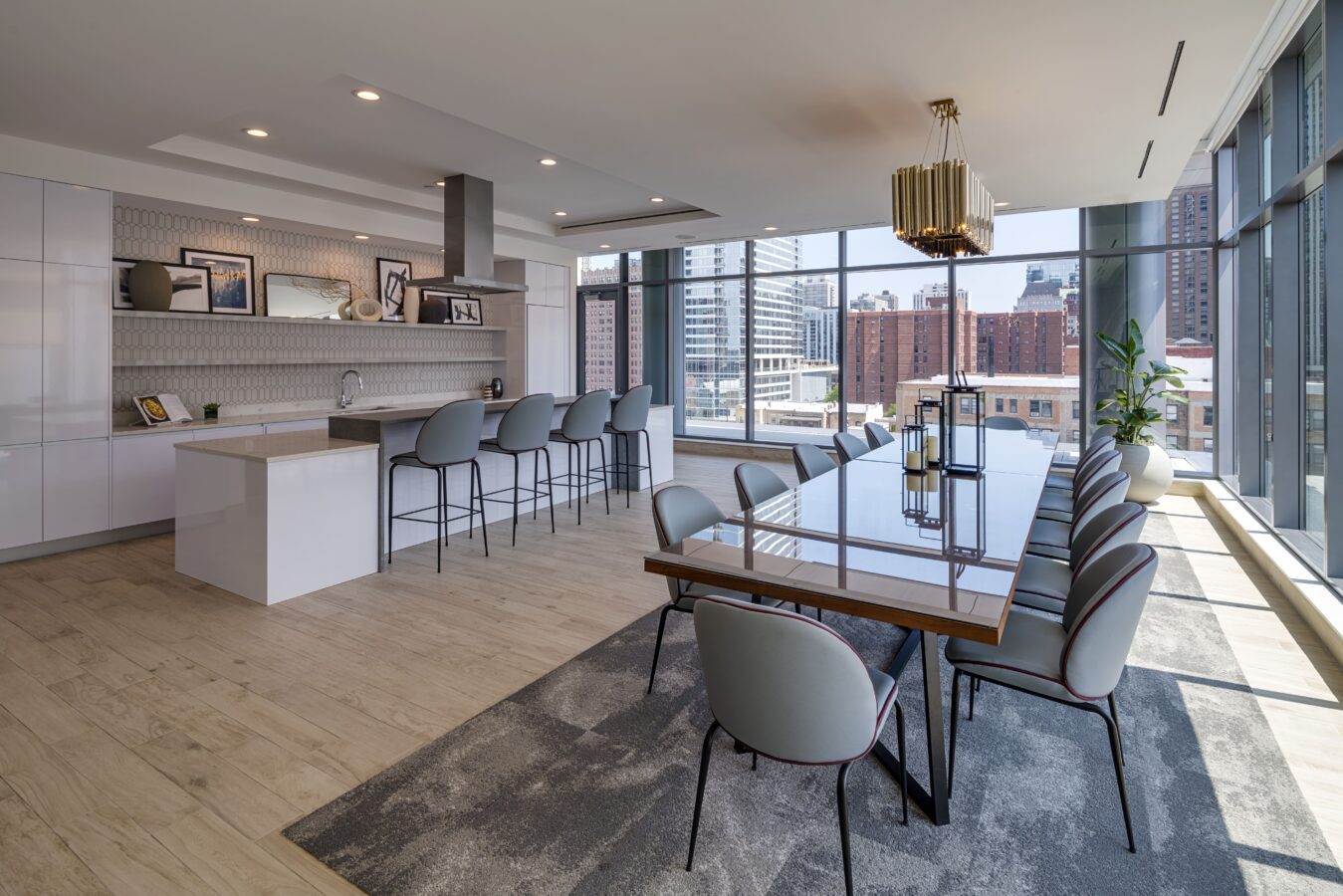 Modern open-plan kitchen and dining area with large windows, city views, a long dining table with gray chairs, a breakfast bar with stools, and stylish lighting fixtures. Light wood flooring and minimalist decor.