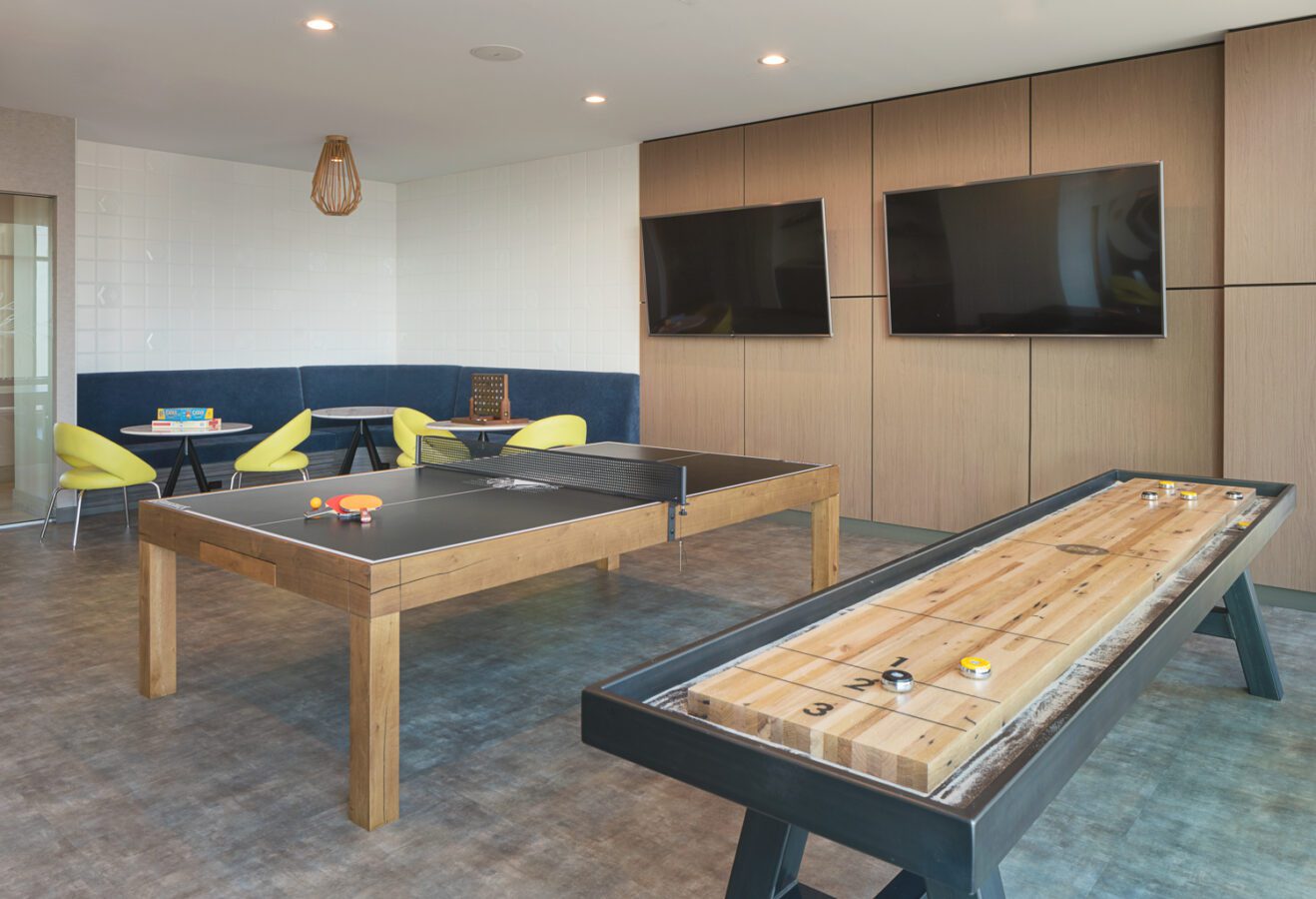 Modern game room with a ping pong table, a shuffleboard table, two wall-mounted TVs, and a seating area with yellow chairs and a small table in the corner. The room has wood paneling and neutral-toned flooring.