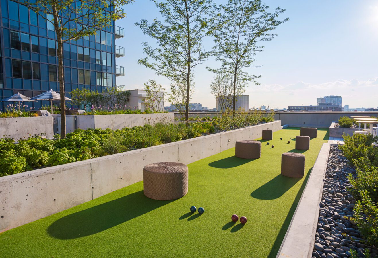 A rooftop bocce court with scattered balls and round woven stools, surrounded by greenery and modern buildings under a clear blue sky.