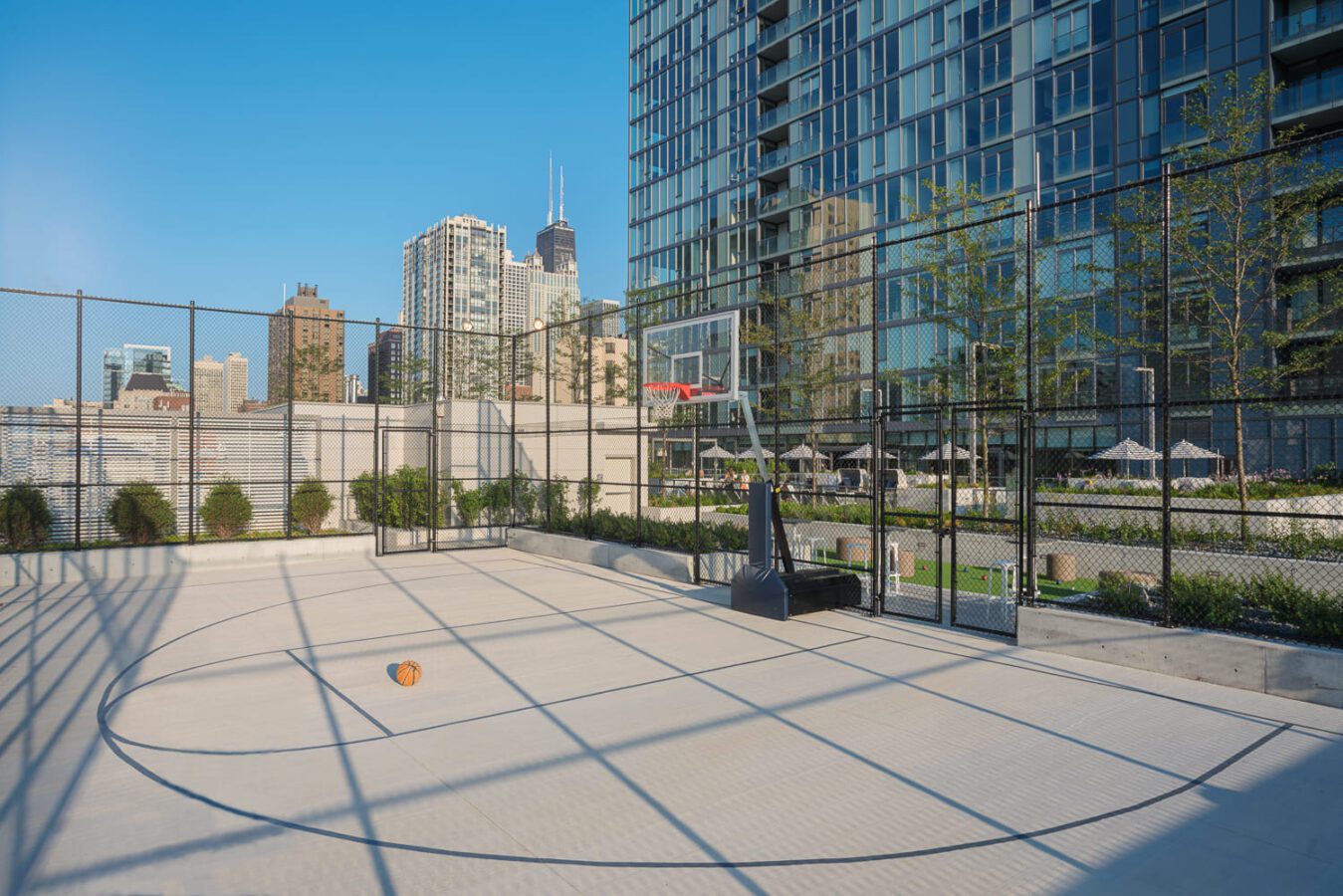 A basketball sits on an empty outdoor court surrounded by a tall black fence, with modern high-rise buildings and a clear blue sky in the background.