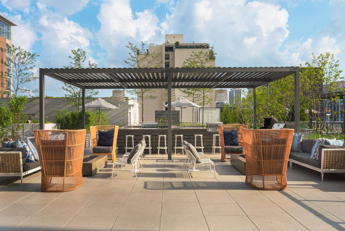 A modern rooftop lounge with shaded seating, orange mesh chairs, cushioned benches, small white tables, and bar stools. Greenery surrounds the space, and buildings are visible in the background under a partly cloudy sky.