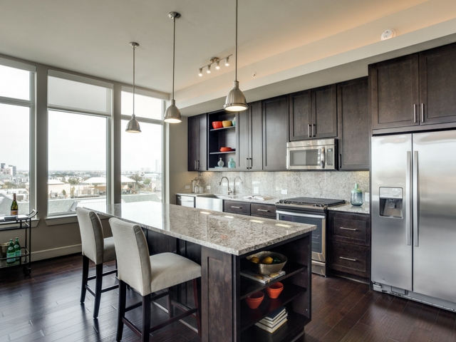 Modern kitchen with dark wood cabinets, stainless steel appliances, granite island with two beige upholstered stools, pendant lights above, and large windows providing ample natural light and a city view.