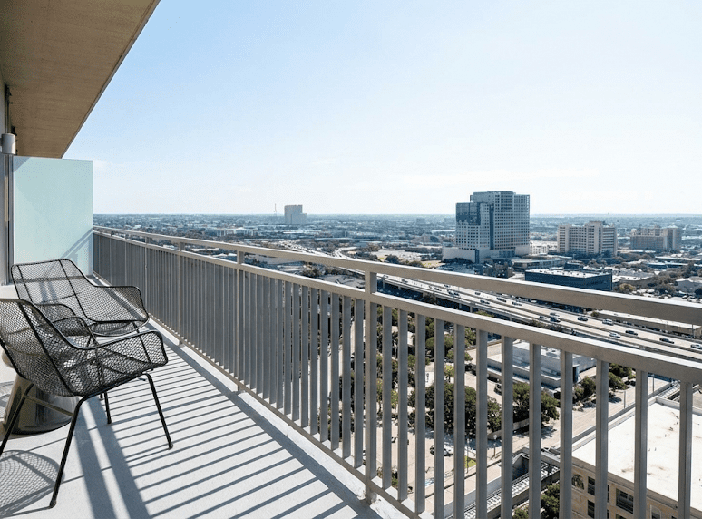 A high-rise balcony with metal chairs and a railing overlooks a cityscape with tall buildings, busy roads, and a clear blue sky in the background.