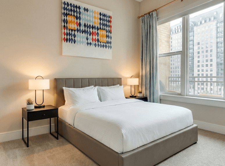 A modern bedroom with a neatly made bed, two bedside tables with lamps, abstract wall art above the headboard, light curtains, and a large window showing city buildings outside.